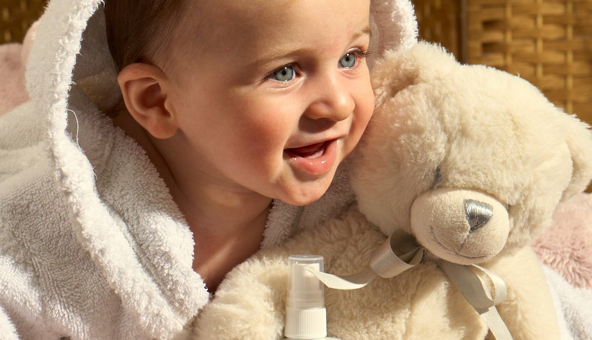 A baby wrapped in a white hooded towel blanket and pink blanket, holding a cream teddy bear and a bottle of Chic Care Soothing Baby Spray.