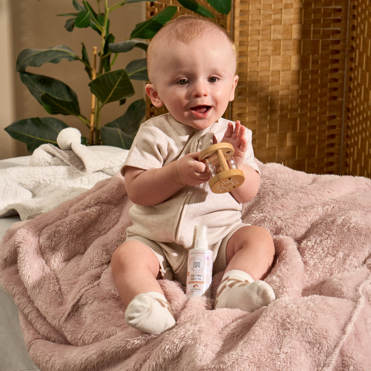 A baby sitting on a pink blanket holding a wooden teething toy, with a small bottle in front of them in a cozy indoor setting.