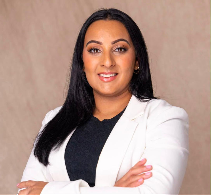 A woman with long black hair stands with arms crossed, wearing a white blazer over a black top, posed against a neutral beige textured backdrop.